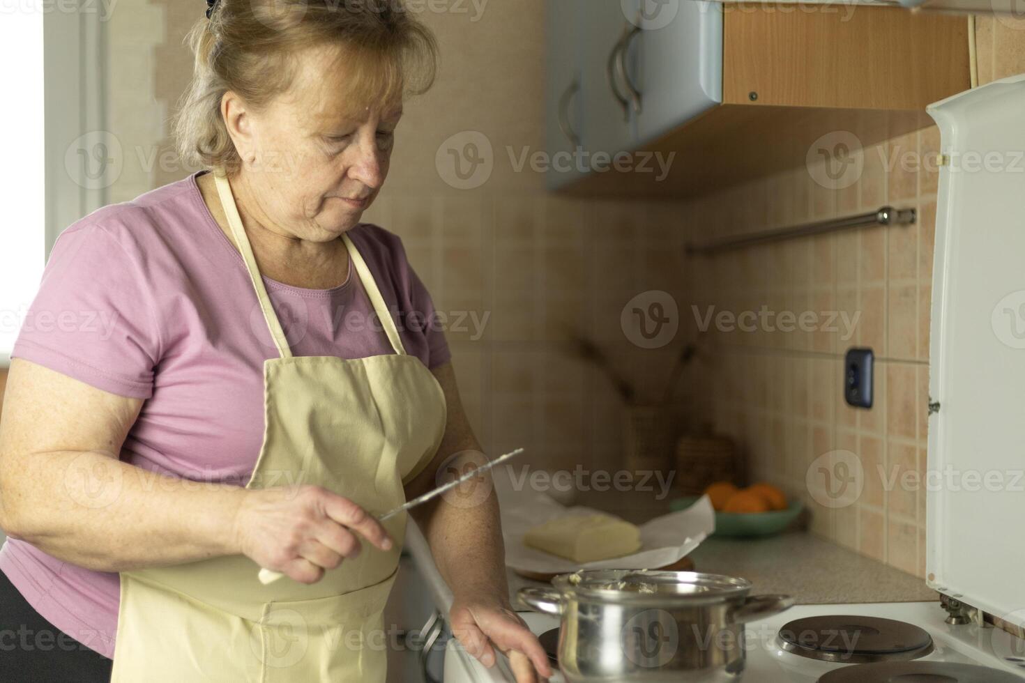 A senior woman in an apron is using a knife to add butter to a saucepan on the stove. photo
