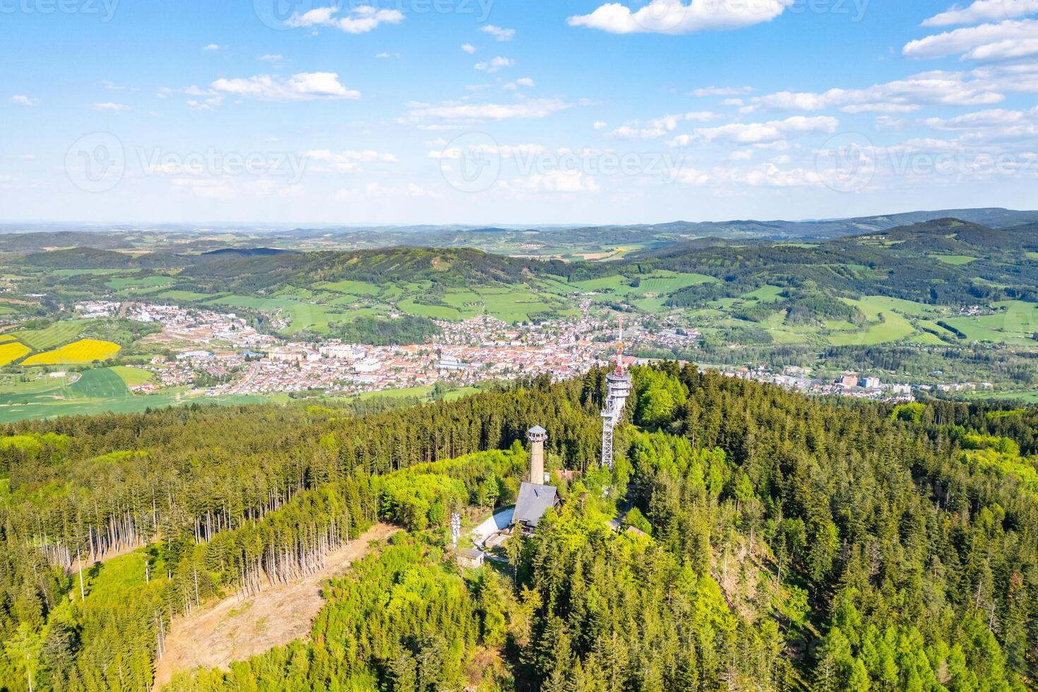 Aerial view captures the Svatobor Lookout Tower surrounded by lush green forests, overlooking the town of Susice nestled in the valley below on a clear day. photo