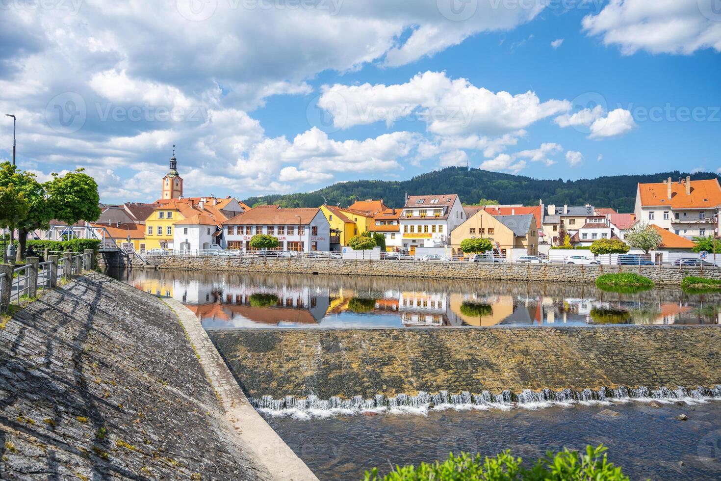 The tranquil waters of the Otava River reflect colorful buildings in Susice, Czechia. Visitors enjoy a sunny day with a backdrop of green hills and blue skies. photo