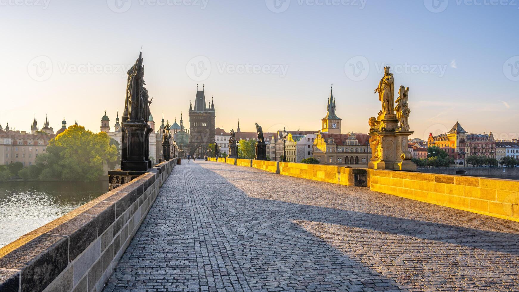 Early morning light casts a warm glow on Charles Bridge in Prague. Statues line the path, while the city awakens with historical buildings reflected in the calm waters of the Vltava River. photo