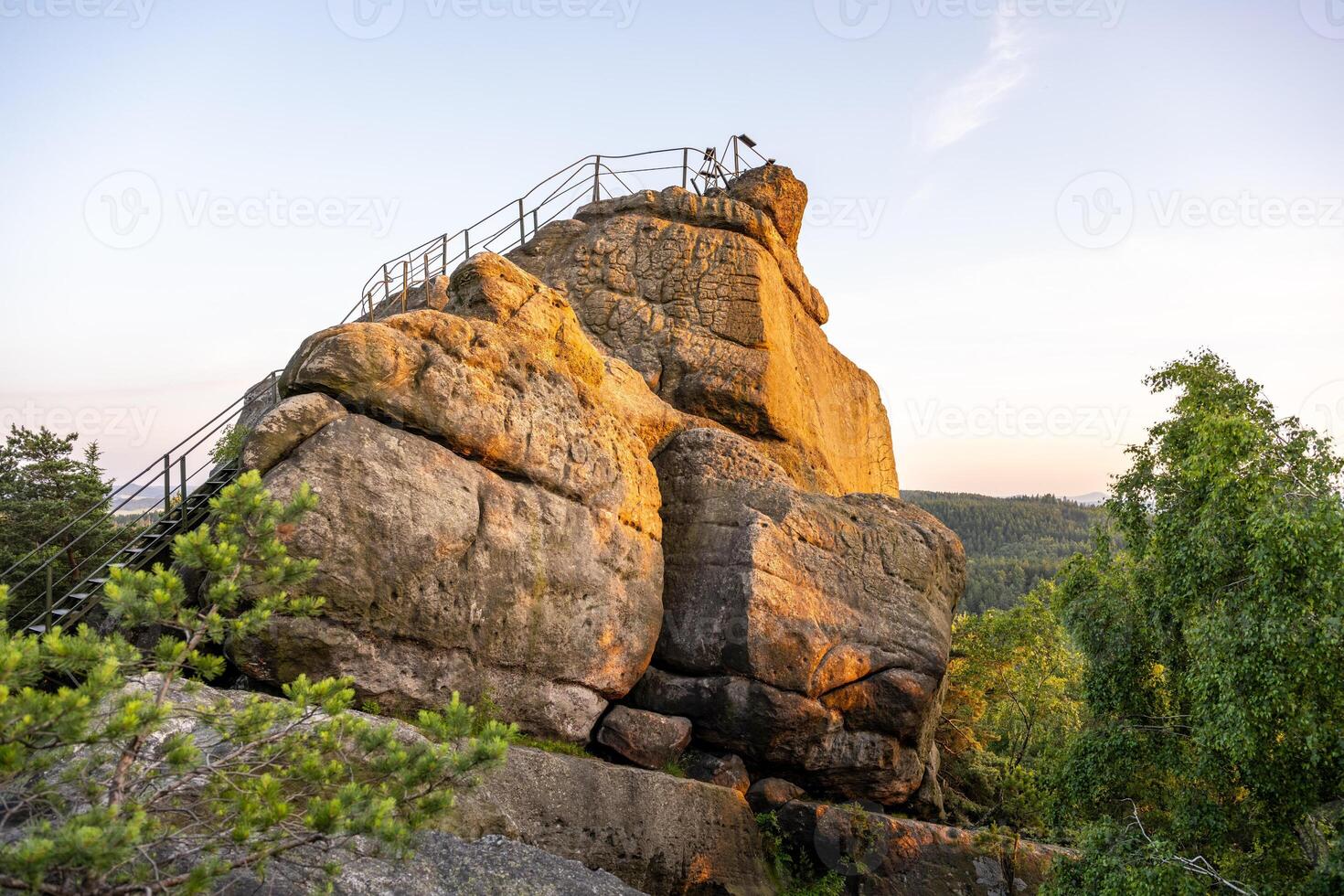 As the sun sets over the Lusatian Mountains, the sandstone Popov Cliffs glow warmly. Visitors can enjoy the beautiful views and explore the rocky terrain during this tranquil evening. photo