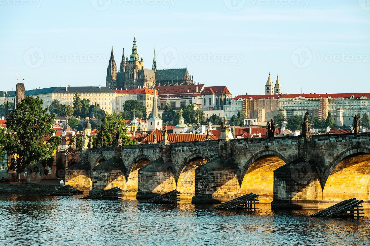 Early morning light illuminates Charles Bridge with Prague Castle in the background. The peaceful Vltava River reflects the stunning architecture, showcasing a tranquil urban landscape. photo