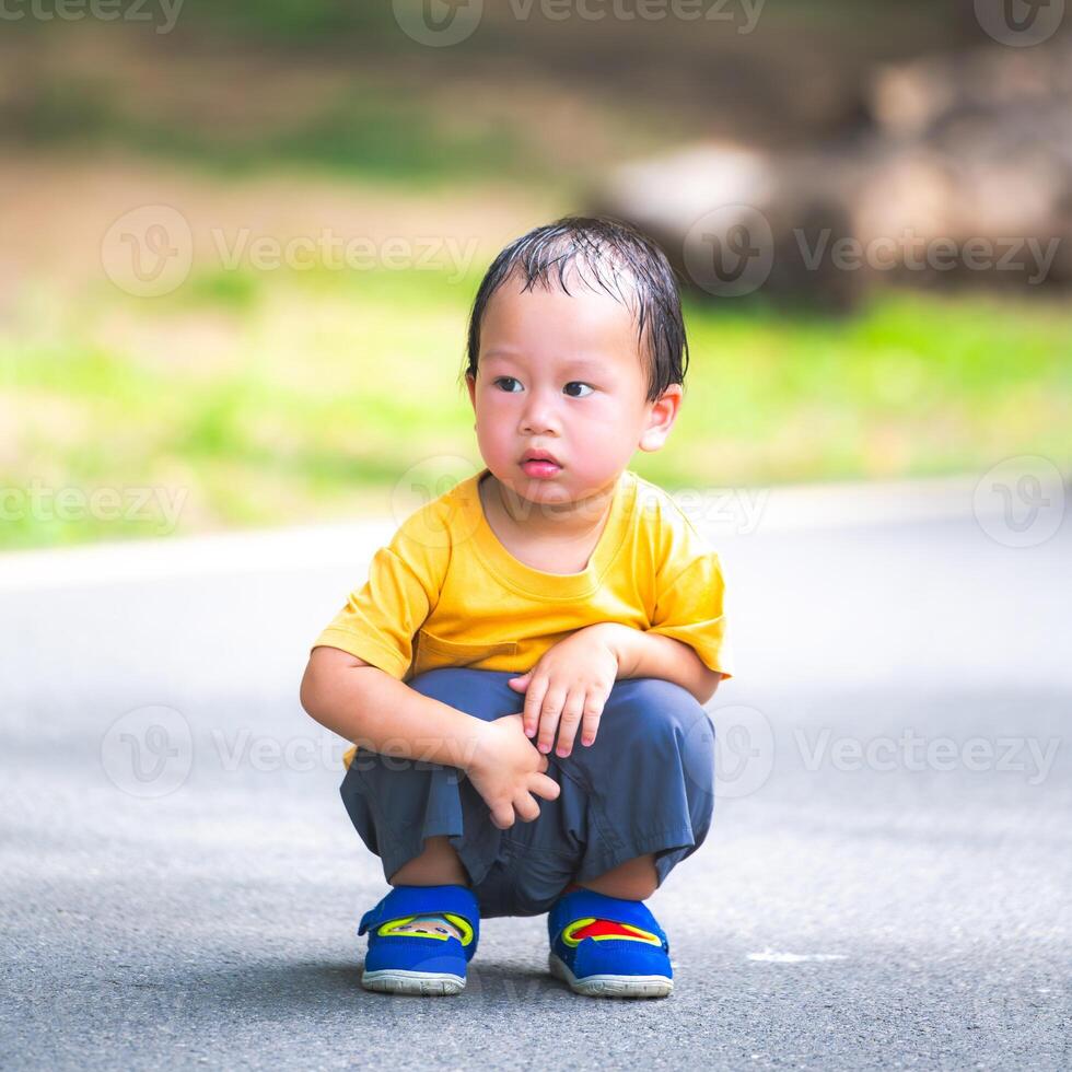 Asian boy's head is soaked in sweat from the hot and humid weather. Child squats in the middle of the road to rest while walking. The 2-year-old kid is wearing a dark yellow shirt. photo