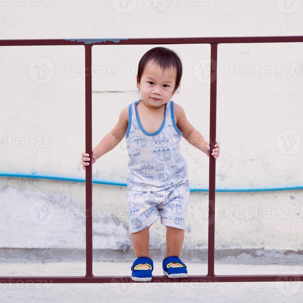 Asian toddler boy having fun climbing on iron fence, toddler happiness, muscle development, happy kid smiling and looking at camera. 2 year old son. photo