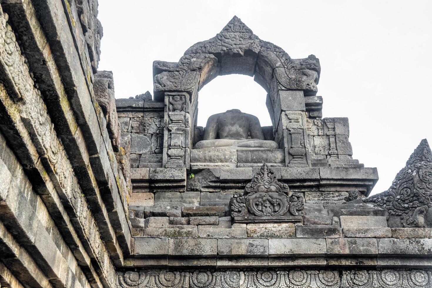 A headless Buddha statue sits in a carved stone arch atop an ancient temple wall. photo