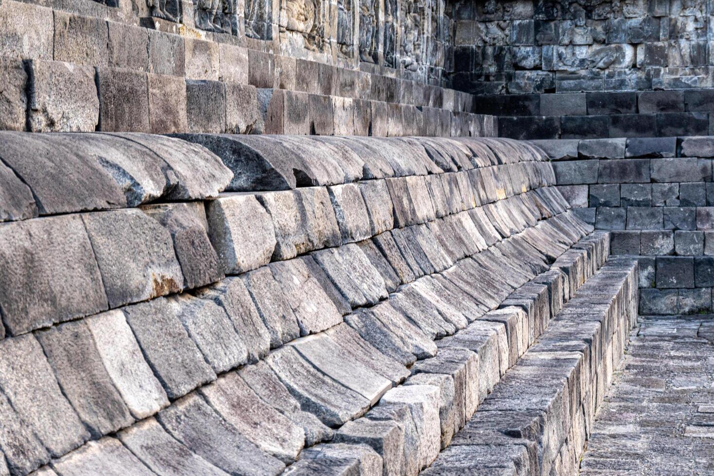 Stone seating structure at Borobudur Temple with worn, curved blocks and ancient carvings above. photo