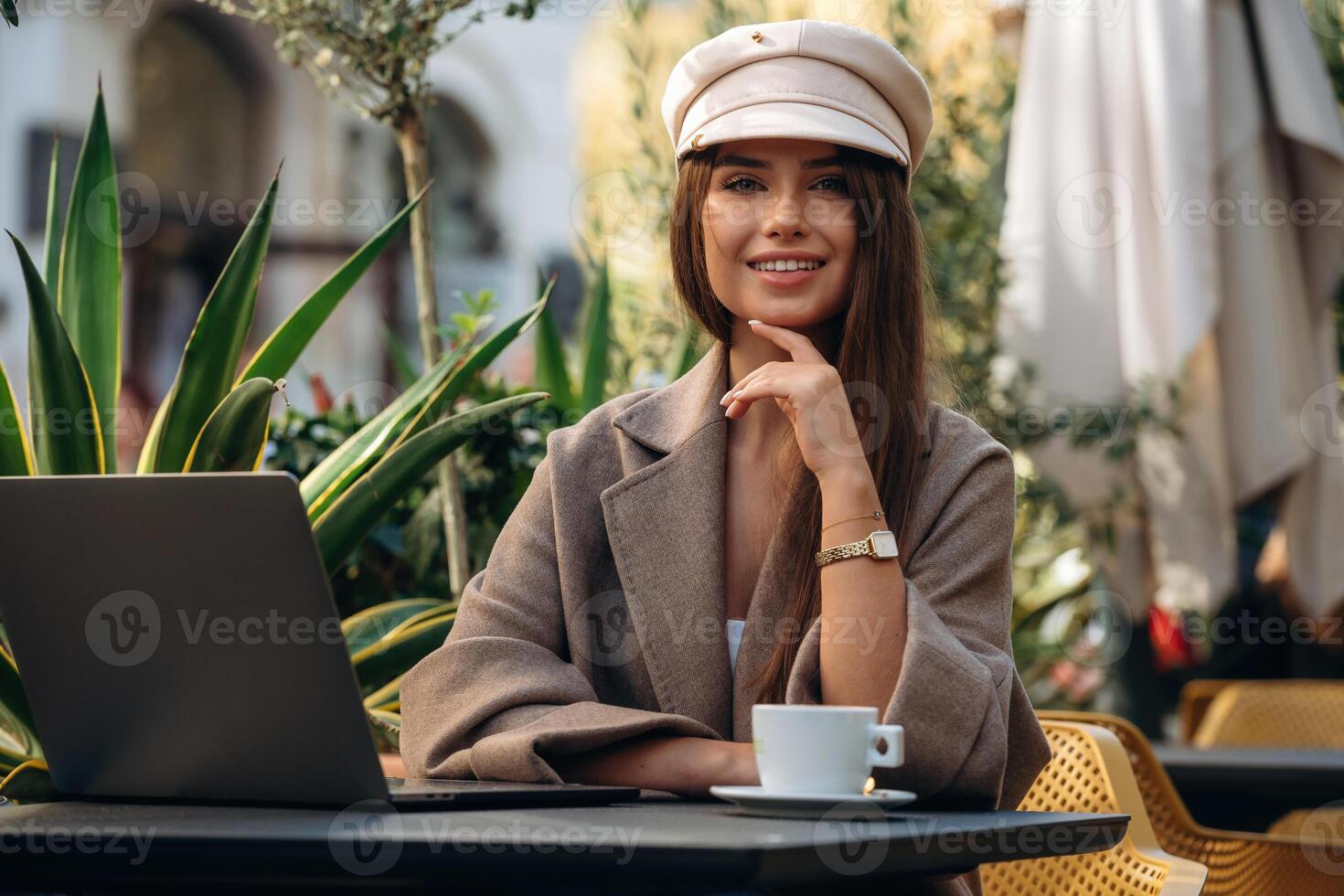 Looking, at the camera, sitting. Beautiful young woman is outdoors of the restaurant by the table photo
