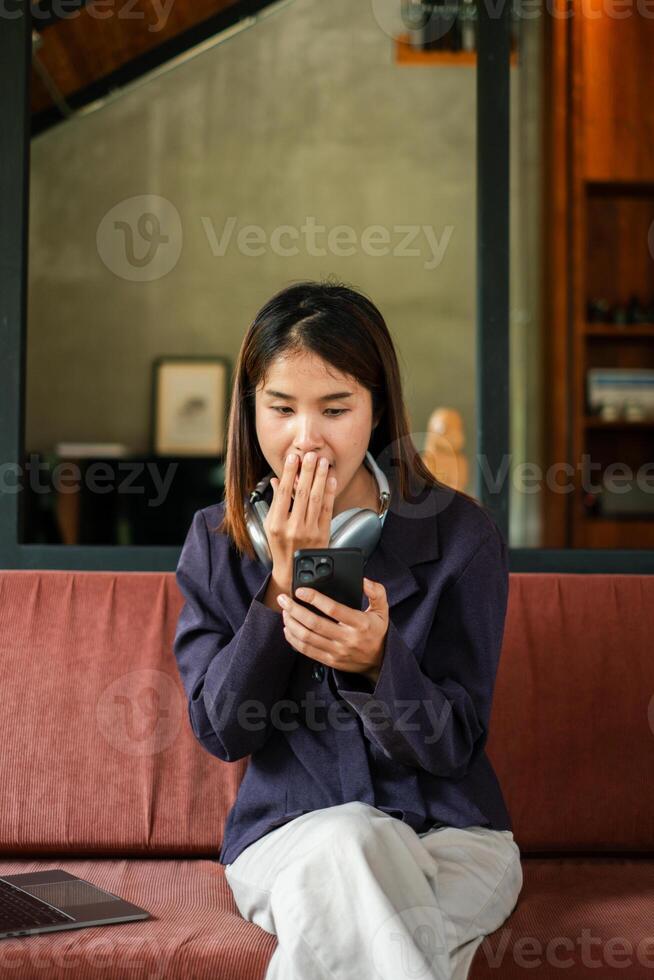 A young woman with headphones around her neck reacts to something on her smartphone while sitting on a sofa in a contemporary living room. photo