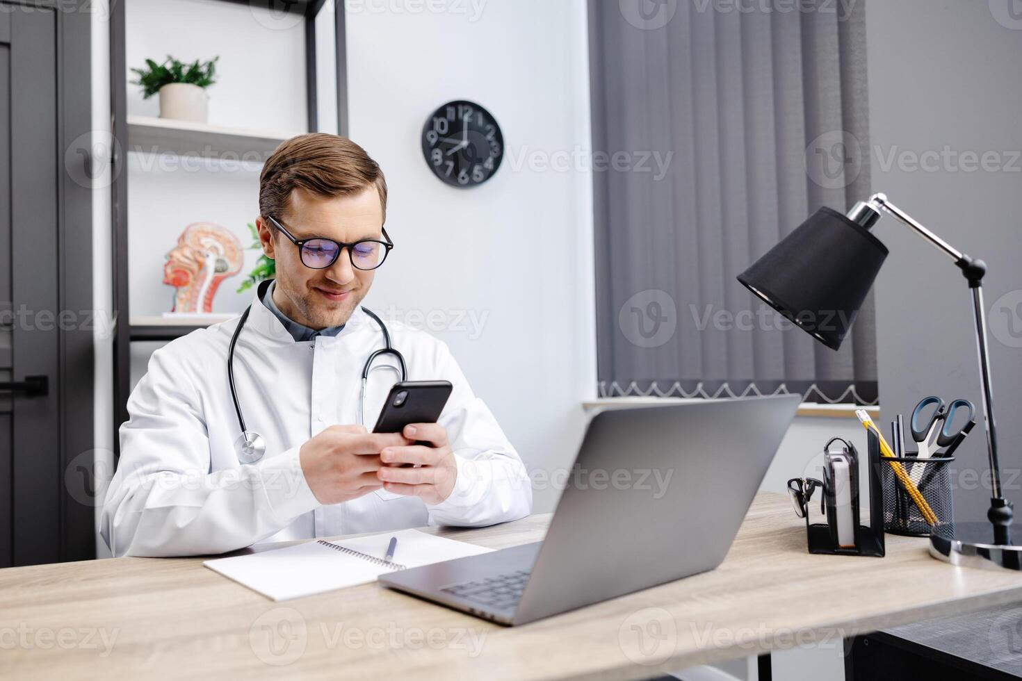 Male doctor medical worker in modern clinic wearing eyeglasses and white coat uniform using cell mobile smartphone apps, sitting at laptop computer photo