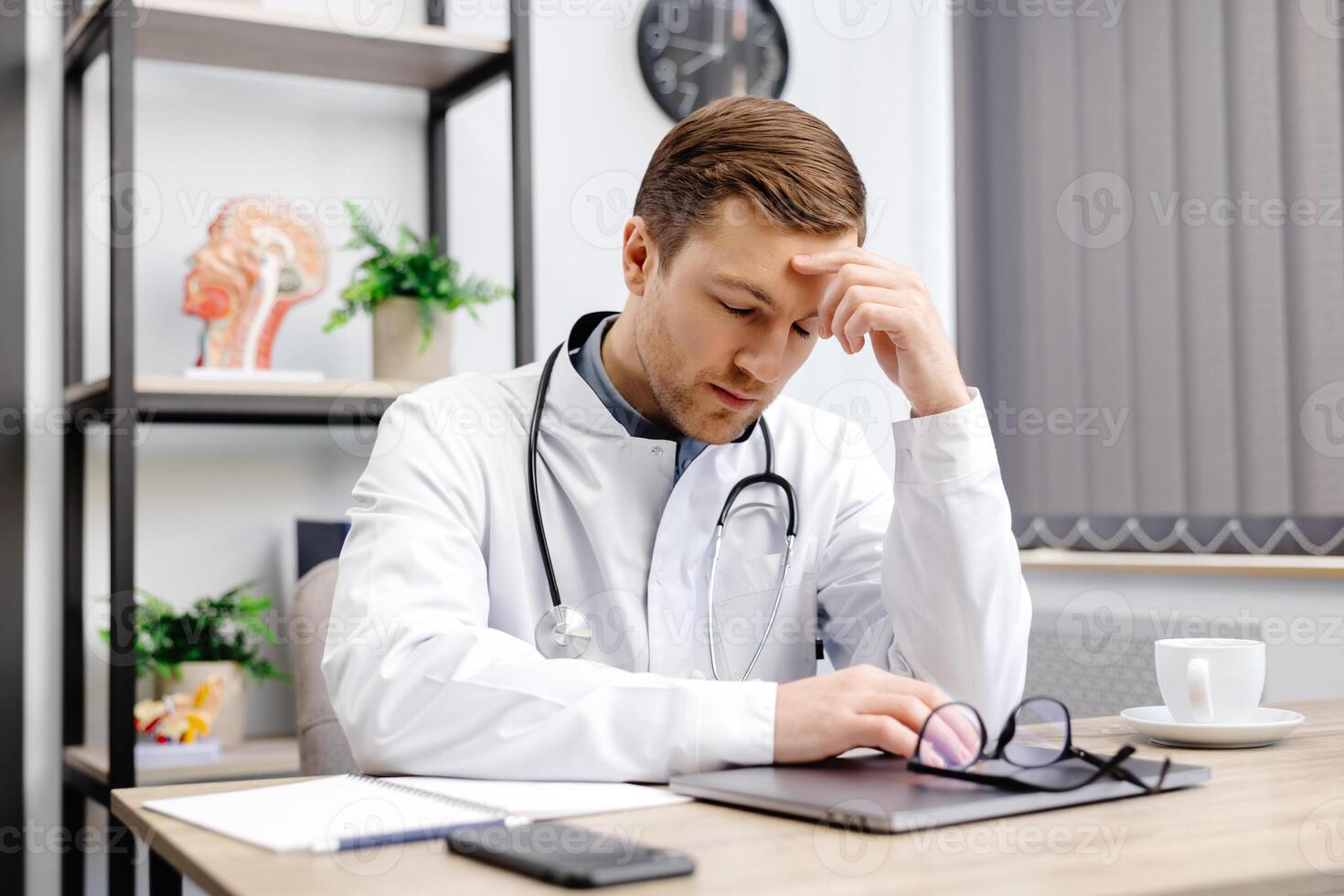 Handsome doctor man wearing medical uniform sitting on his workplace tired holding his head feeling fatigue and headache. Stress and frustration concept. photo