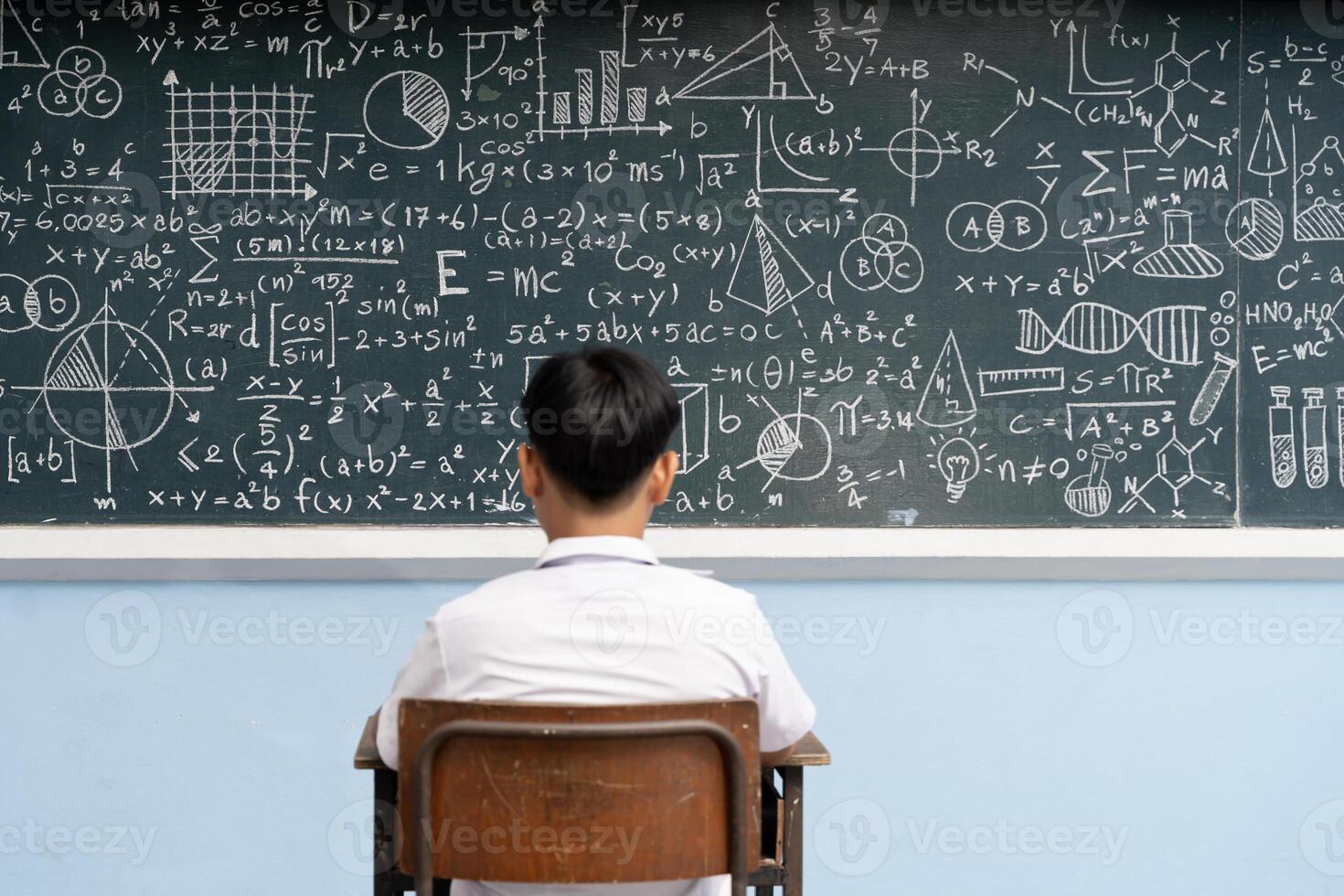 Student sitting alone facing blackboard full of complex math formulas, equations, geometry, science drawings in classroom photo