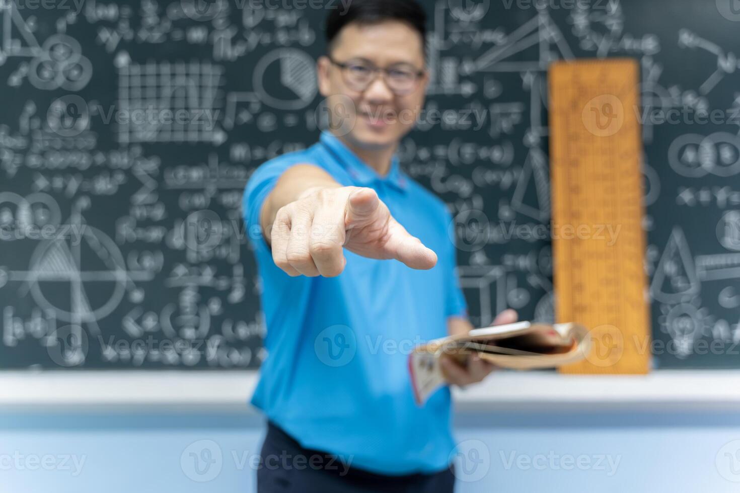 Asian math teacher pointing directly at camera holding book standing in classroom with blackboard of formulas and geometry photo