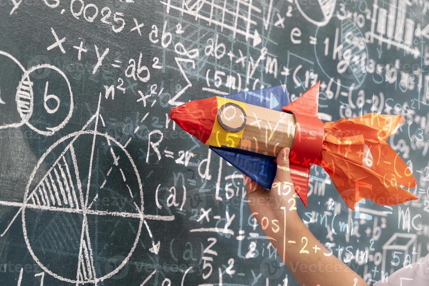 Hand holding colorful rocket craft in front of blackboard full of math and science formulas with overlay effect photo