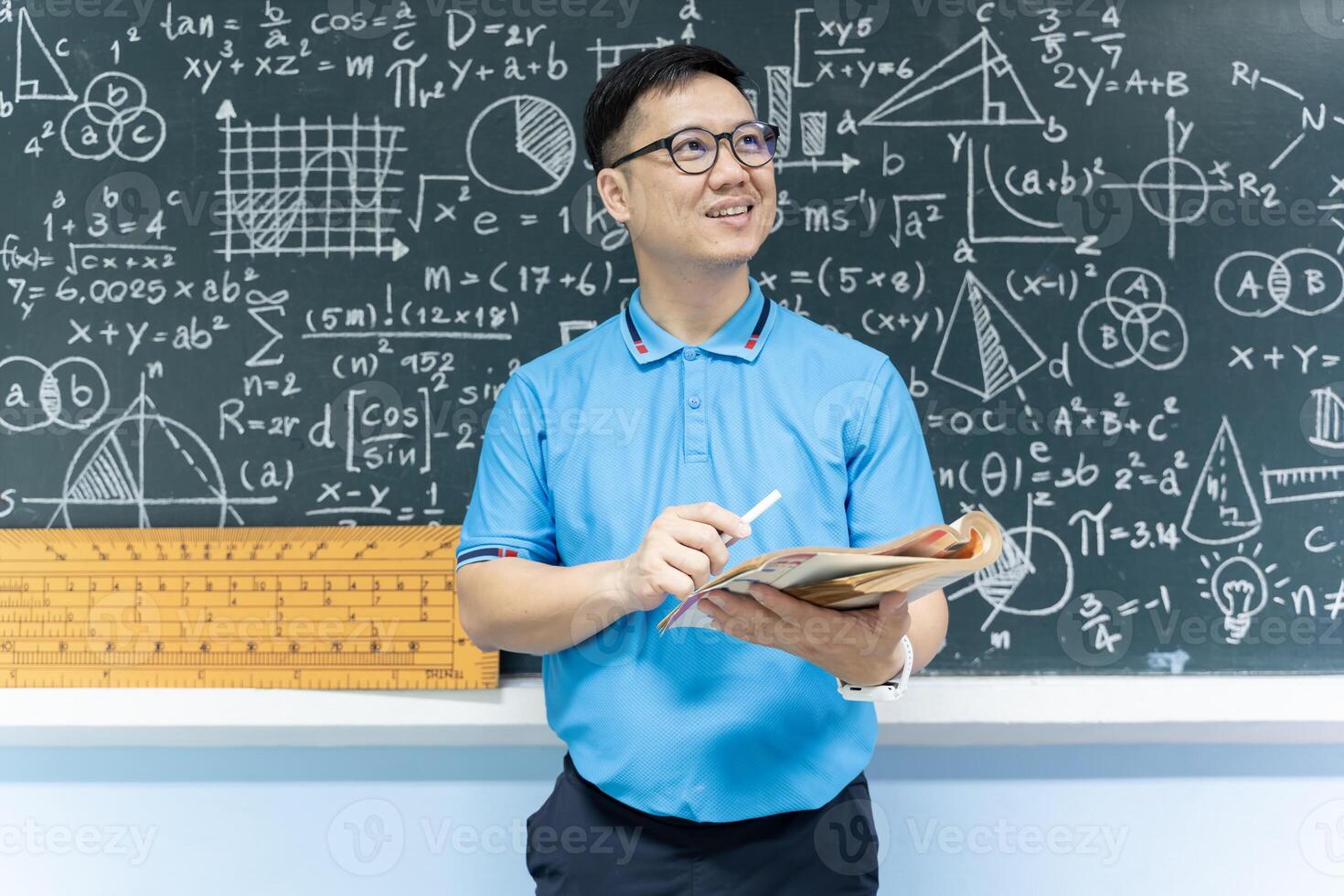 Asian male teacher holding book and chalk standing in classroom with blackboard full of math formulas and geometric drawings photo