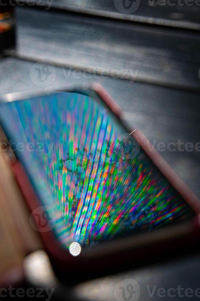 Bright rainbow rays reflect in mobile phone screen surface with dark blurred background. Multicolored beams and reflection in smartphone screen closeup photo
