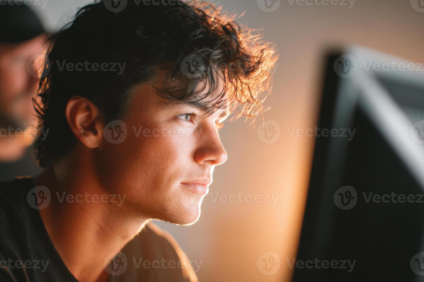 Young man with dark hair is focused on a computer screen, illuminated by warm light, showcasing concentration and creativity in a modern workspace environment photo