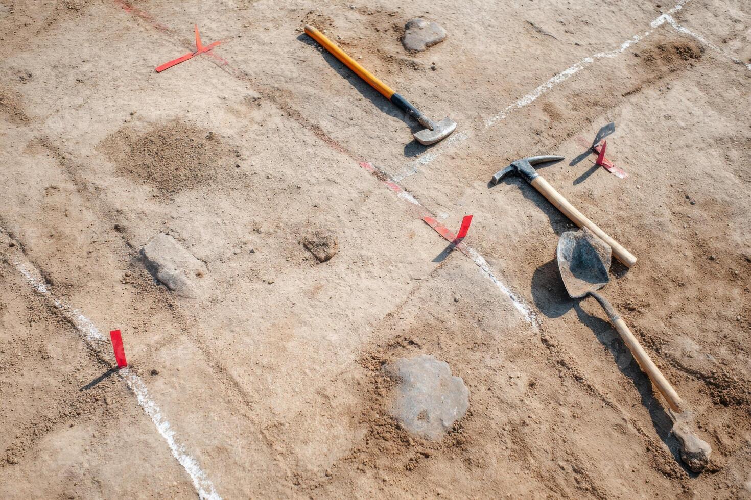 Archaeological excavation site featuring various tools including hammers and shovels on sandy soil, marked with red flags, showcasing the meticulous process of uncovering historical artifacts photo