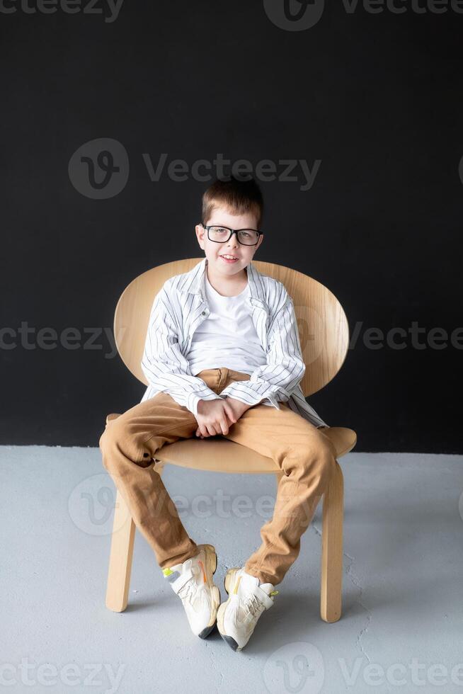 An 11 year old boy with reading glasses and hearing aids in a white shirt sits on a table against a black wall and smiles while looking at the frame photo