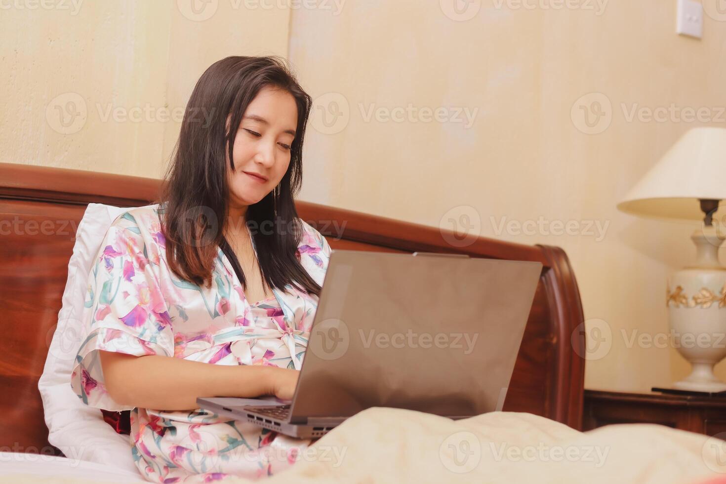 Woman enjoys working on laptop in bed, wearing floral robe for c photo