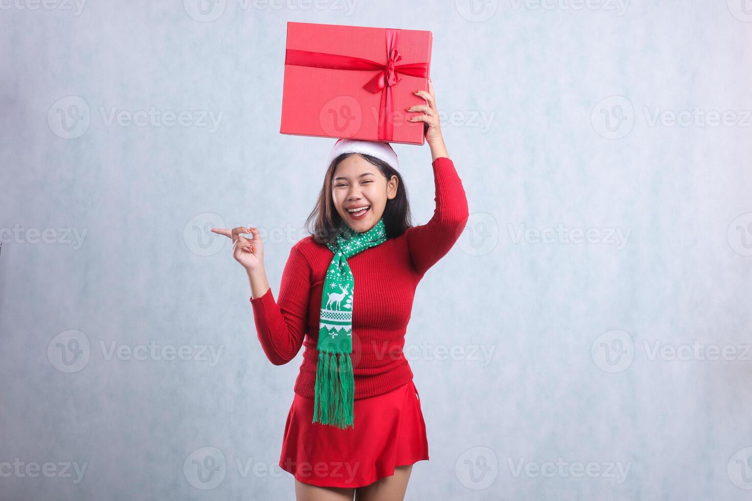 cheerful young asian female looking at camera hand holding red merry christmas box gift and pointing right, wearing christmas sweater party outfit, santa hat and scarf, isolated on white background photo