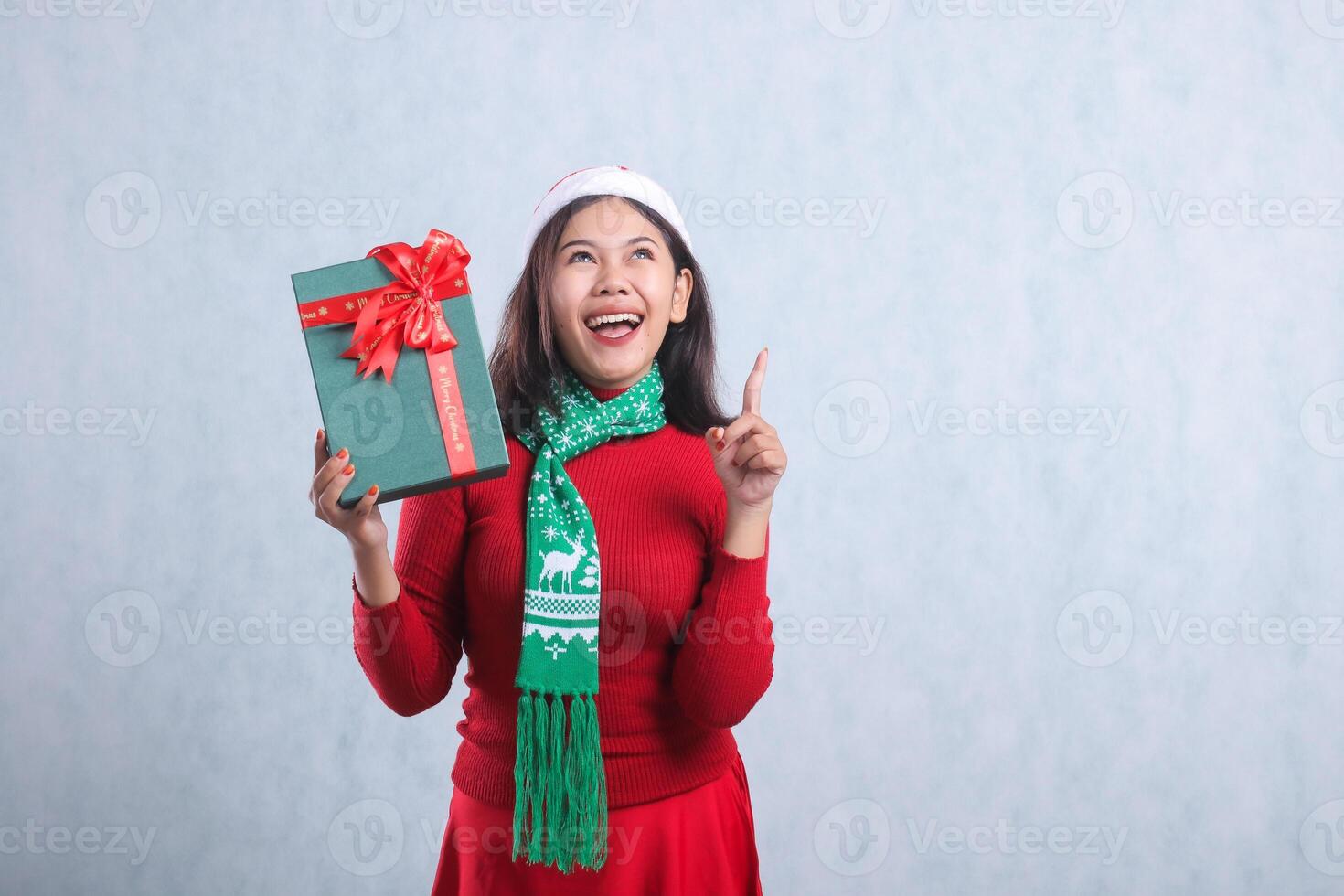 Charming adult woman wearing red christmas sweater, Santa hat and scarf, happy upward hand holding green merry christmas box gift and pointing up, isolated on white background photo