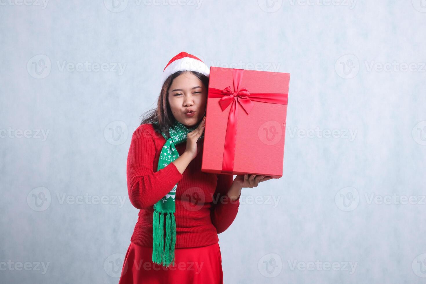 portrait of woman wearing christmas sweater, santa hat and scarf, kissing looking at camera both hands carrying red merry christmas box gift, isolated on white background photo