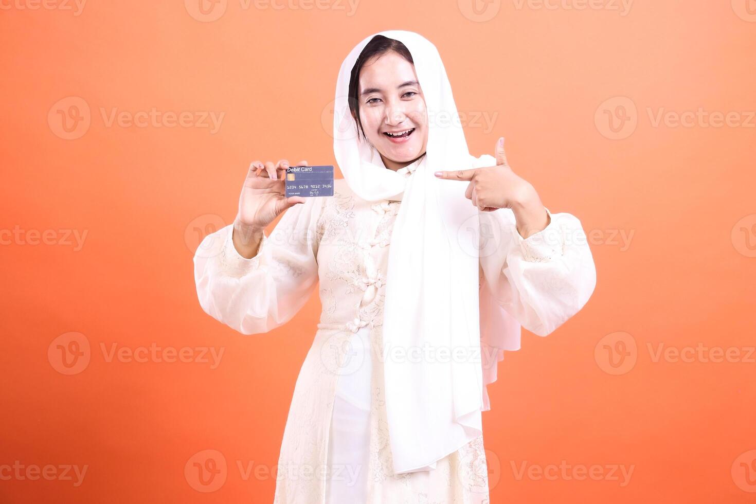 Cheerful Muslim woman showcasing debit card for online payments and banking needs in contemporary finance, isolated on a orange background. photo