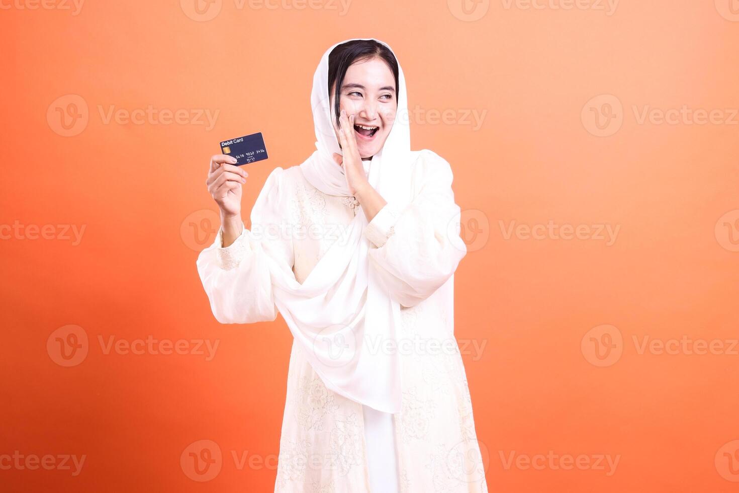 Excited Muslim Woman Holding Credit Card, isolated on a orange background, showcasing financial opportunity and modern banking accessibility. photo