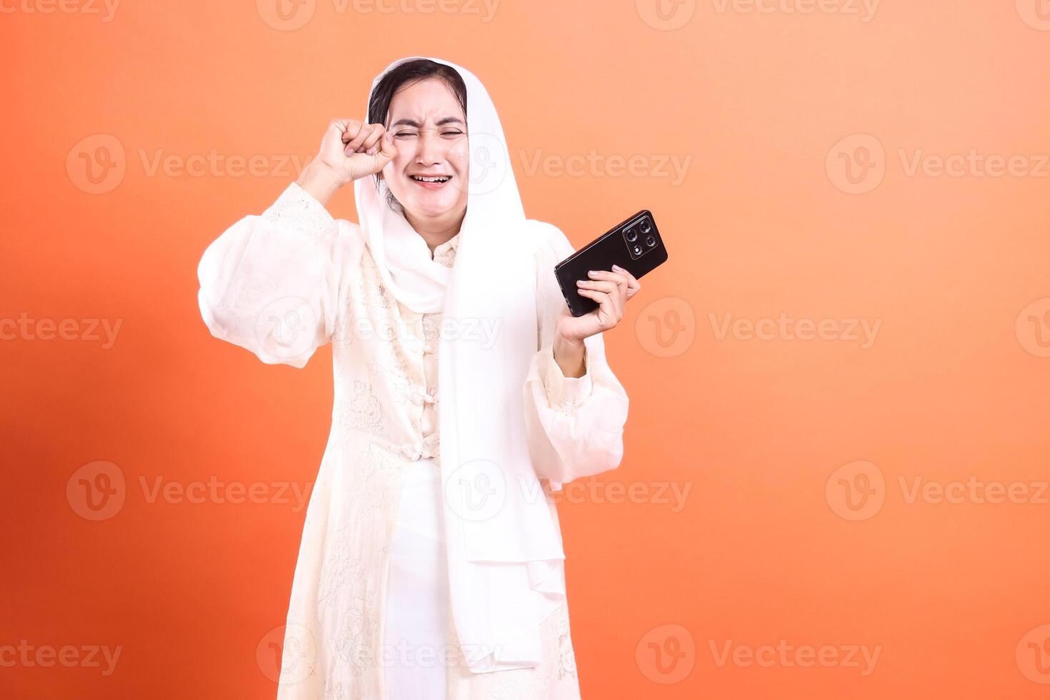 Sad Muslim woman crying after seeing bad news on her phone, wearing a hijab in despair, against a vibrant orange backdrop, in a studio shot. photo