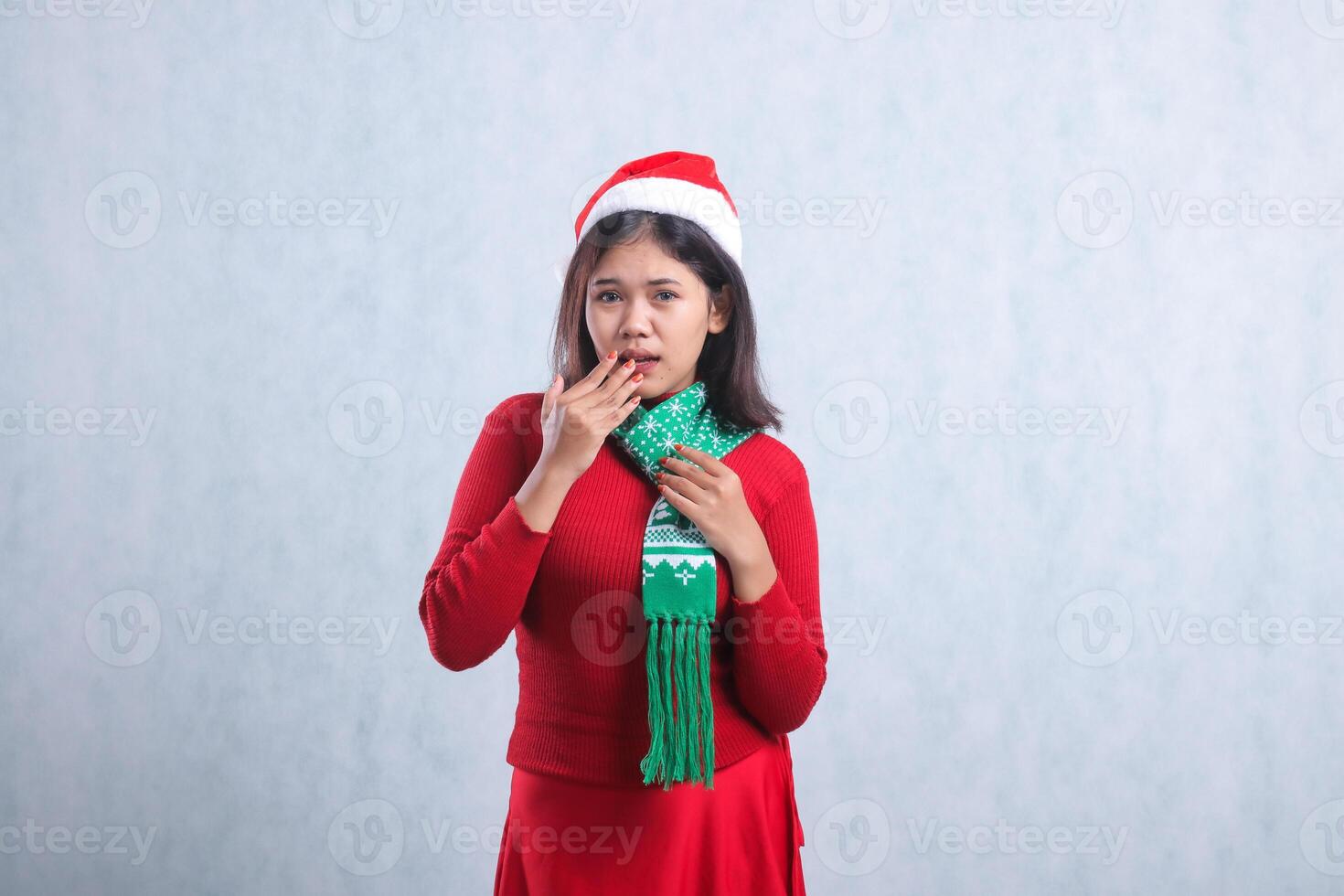 expression of young indonesia woman wearing christmas sweater, santa hat and scarf, afraid to camera hand holding scarf covering mouth, isolated on white background photo