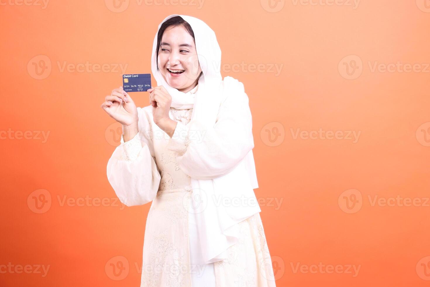 Happy Muslim Woman with Debit Card The Convenience of Modern Finance in Traditional Life showing a credit card isolated on a orange background. photo