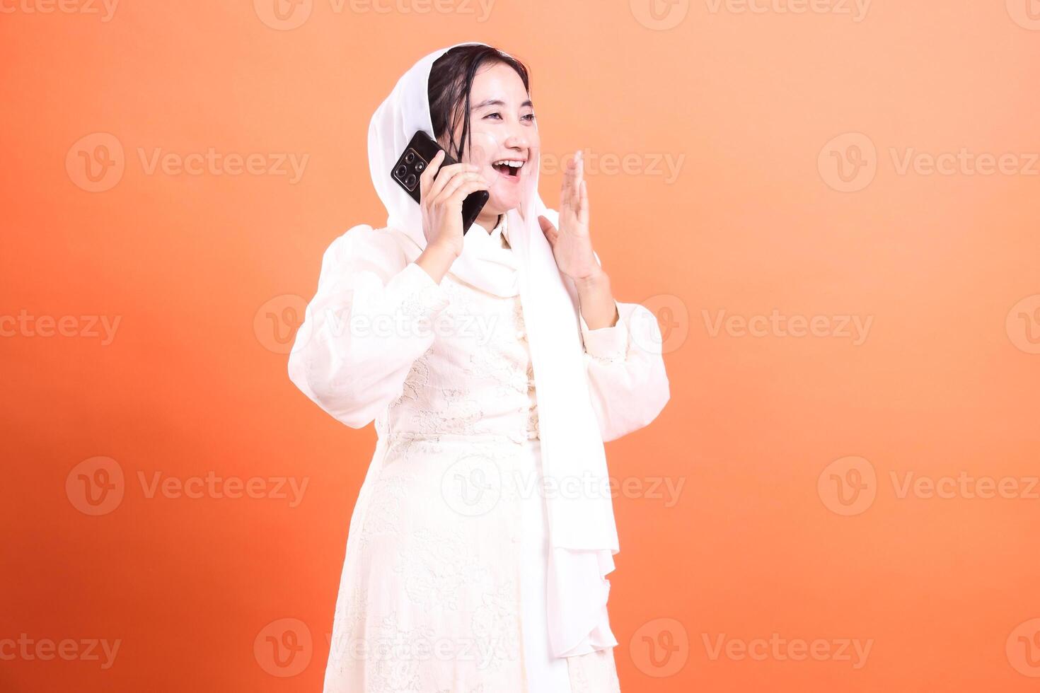 Excited Muslim Woman Chatting on Phone A vibrant image capturing a cheerful woman in hijab with smartphone, radiating joy, isolated on a orange background. photo