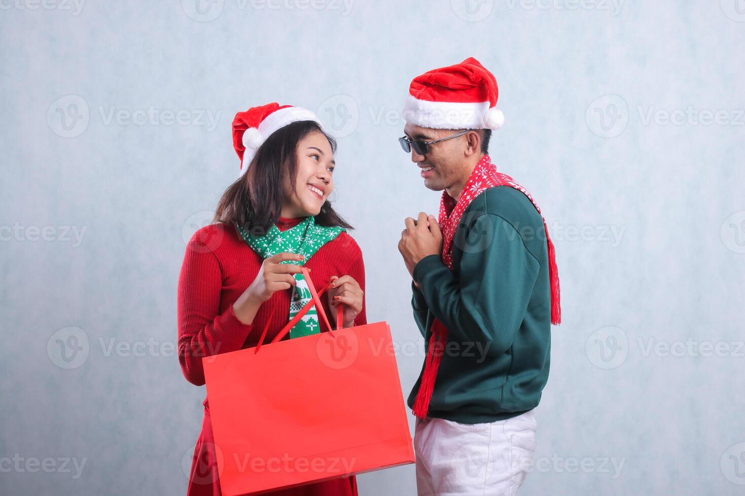 gesture of adult Asian couple wearing Christmas sweater, Santa hat and scarf, happily looking at each other face while carrying Christmas gift paper bag, isolated on white background photo