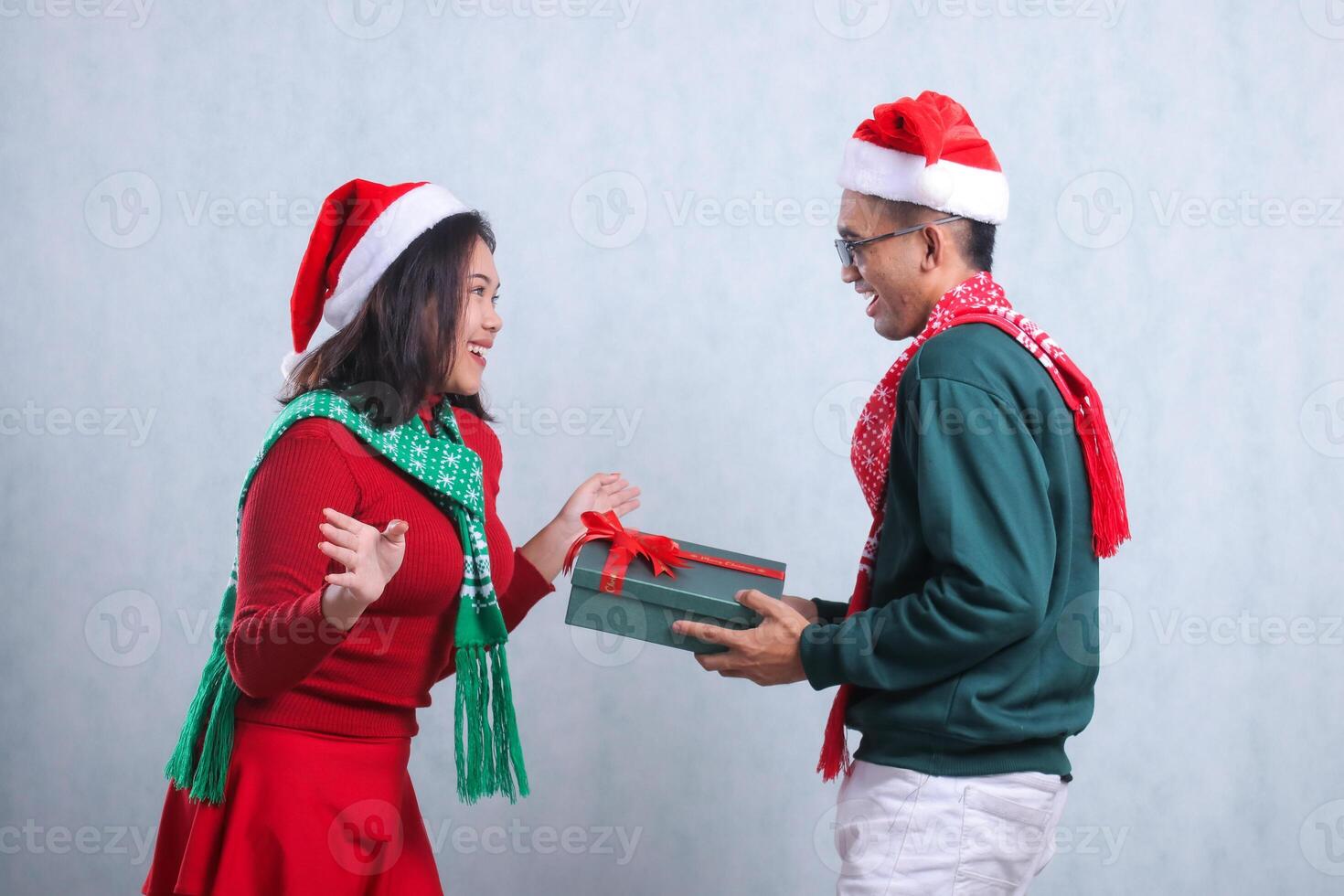 gesture of young couple wearing Christmas sweater, Santa hat, and scarf, cheerfully surprised receiving Christmas gift box from their partner looking at each other, isolated on white background photo