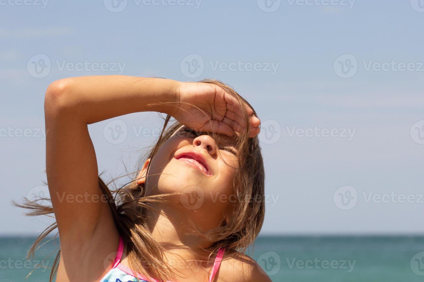 Beautiful caucasian child of 7-8 years looking at sun and covering face with hand on sea background photo