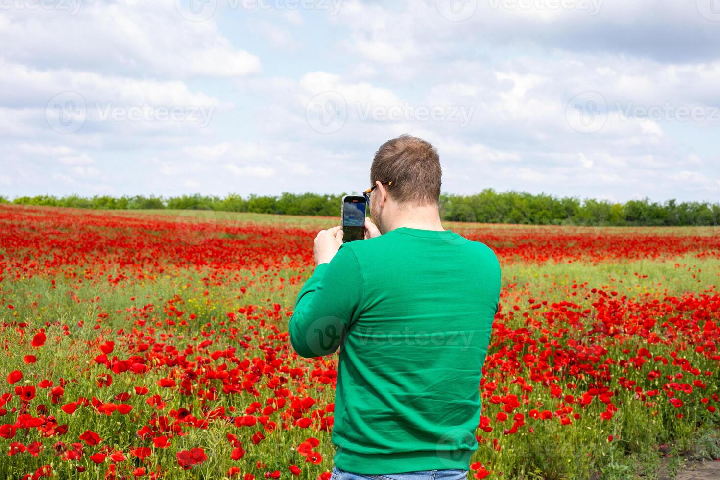 A Caucasian man in a verdant sweater captures the scarlet poppy sea under cotton cloud skies on World Photography Day photo