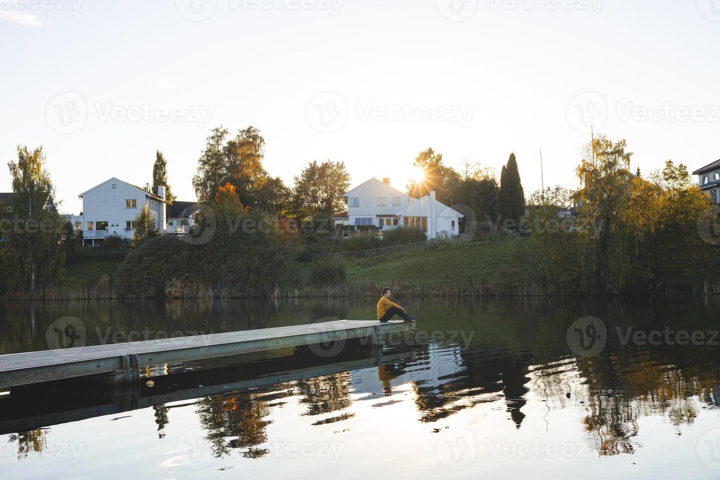 Lone man sits on pier an Oslo lake, Holmendammen as the sun sets behind charming houses, capturing a peaceful moment of reflection and stillness in Norway autumn magic photo