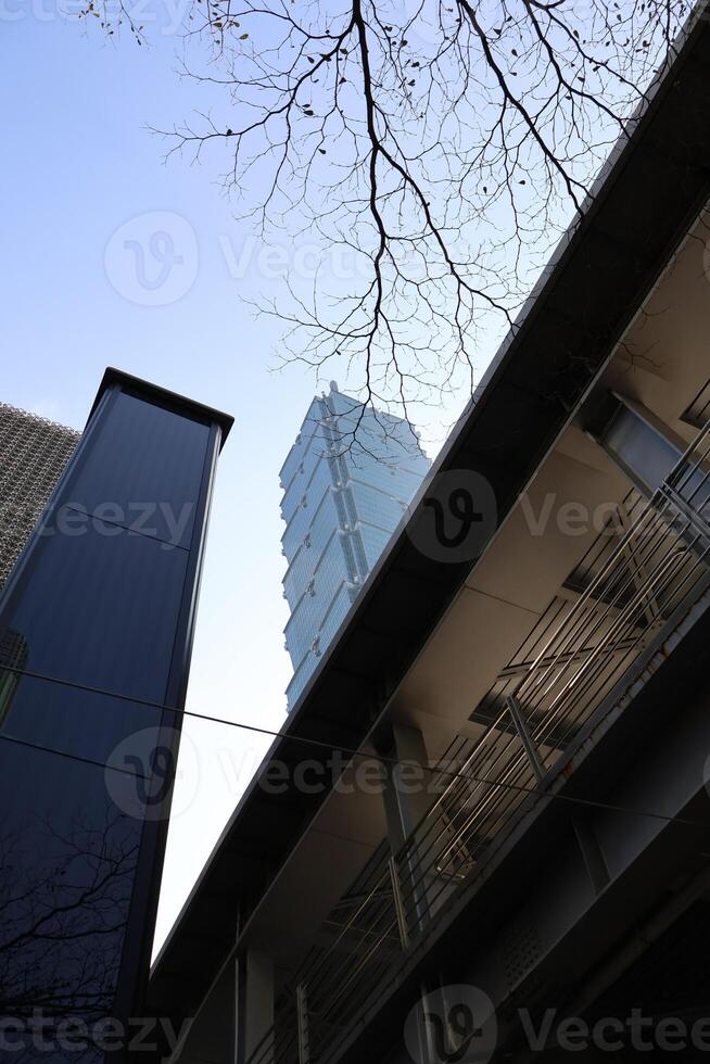 A perspective looking up at Taipei 101 on a clear day, partially obstructed by buildings and trees. photo