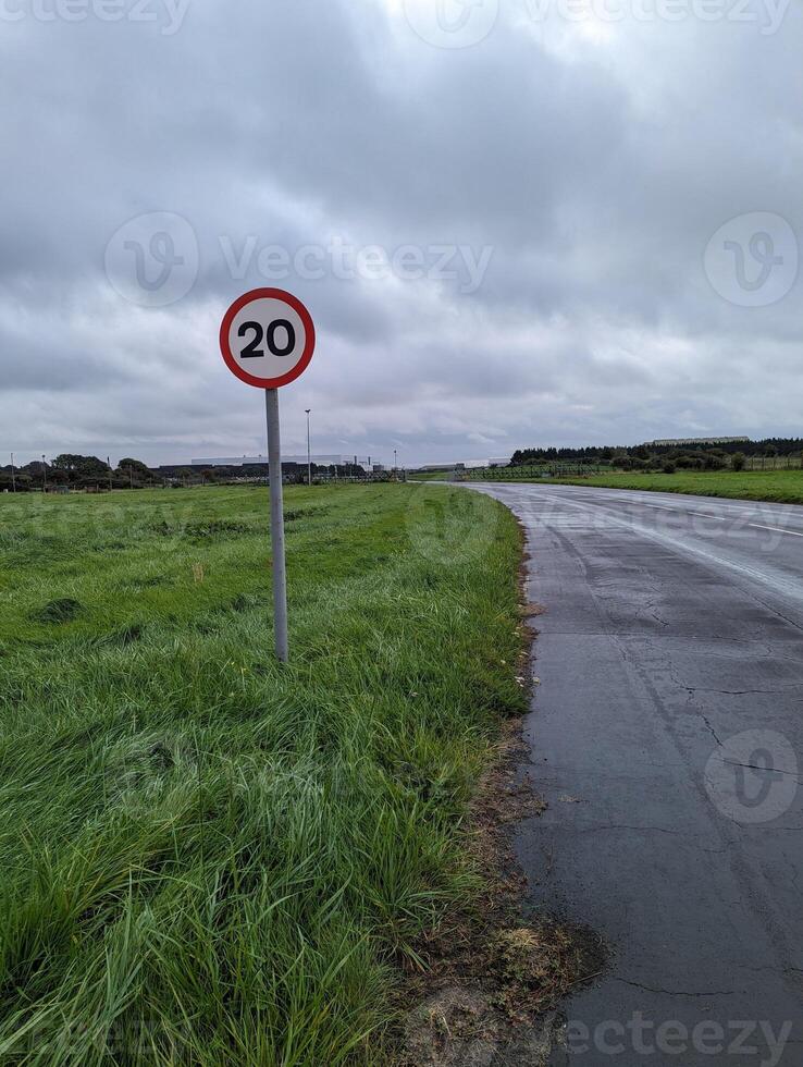 View of 20mph warning sign alongside low trafficked road, surrounded by grass and cloudy sky. photo