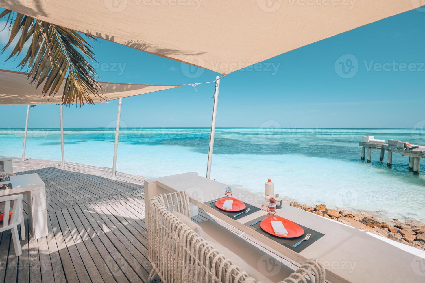 Relaxing outdoor restaurant at the beach. Table setting at tropical beach restaurant. Elegant white wooden tables, chairs under beautiful soft sunset sky, sea view. Luxury hotel or resort restaurant photo