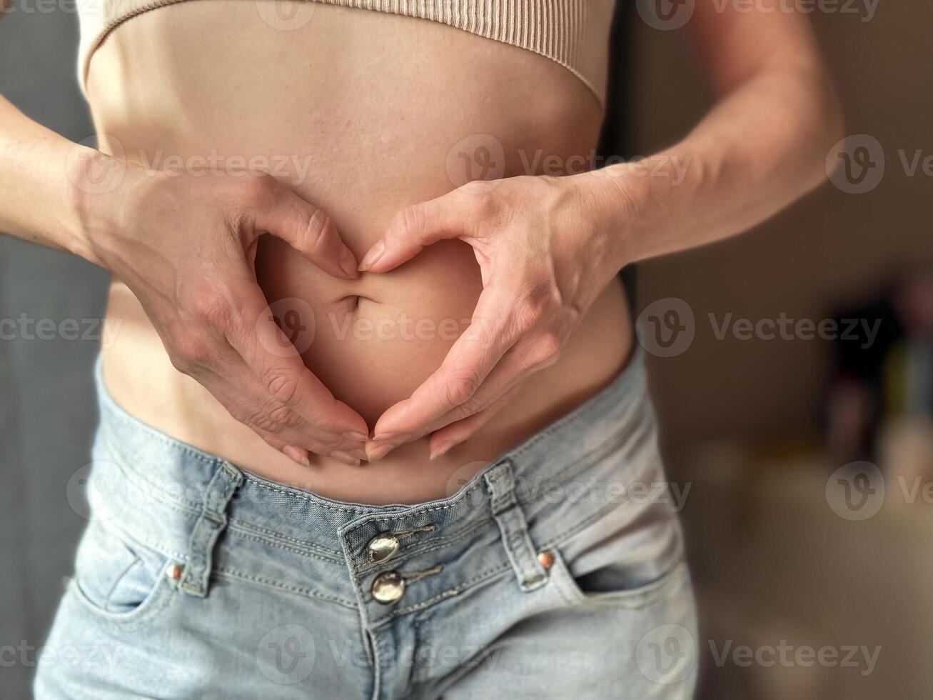 Close up of slim woman in short top and jeans forming heart shape with her hands on her bare stomach. Concept of body positivity, self love, and health awareness. Lifestyle photo. photo