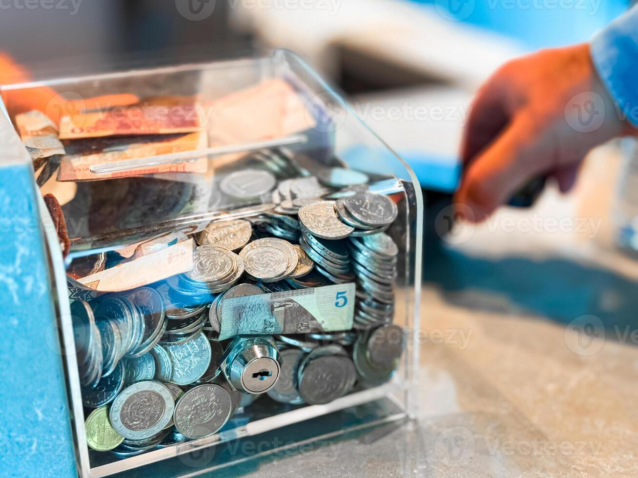 Transparent donation box filled with various coins and banknotes on countertop with hand holding credit card in background. Close up shot for financial aid and charity concept. photo