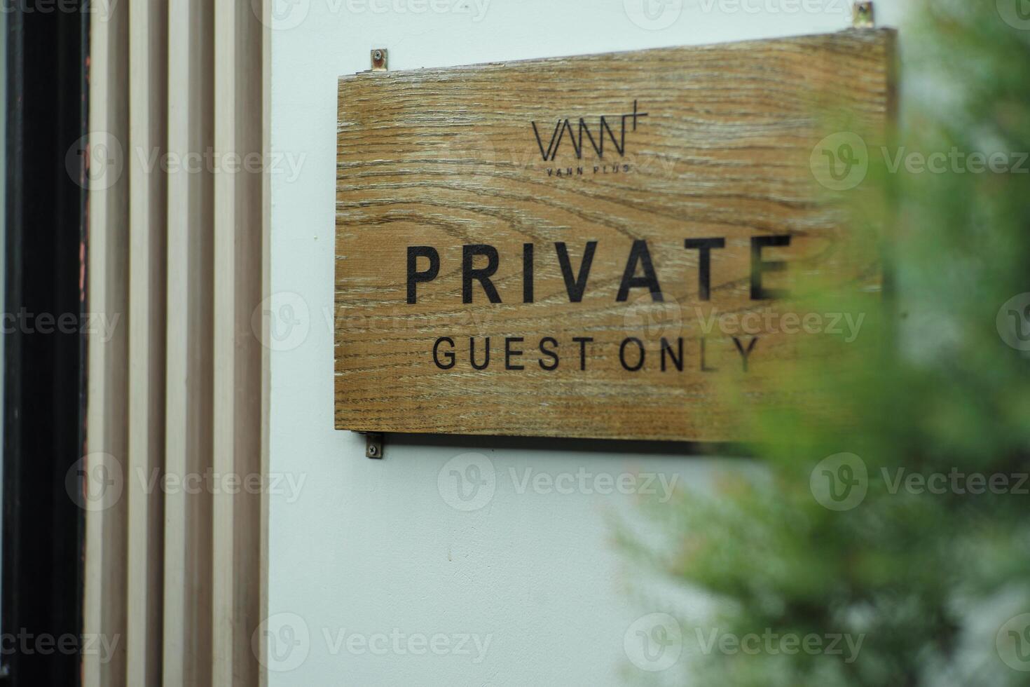 The wooden signboard with the words Private Guest Only hanging on the white wall with blurred tree in foreground photo