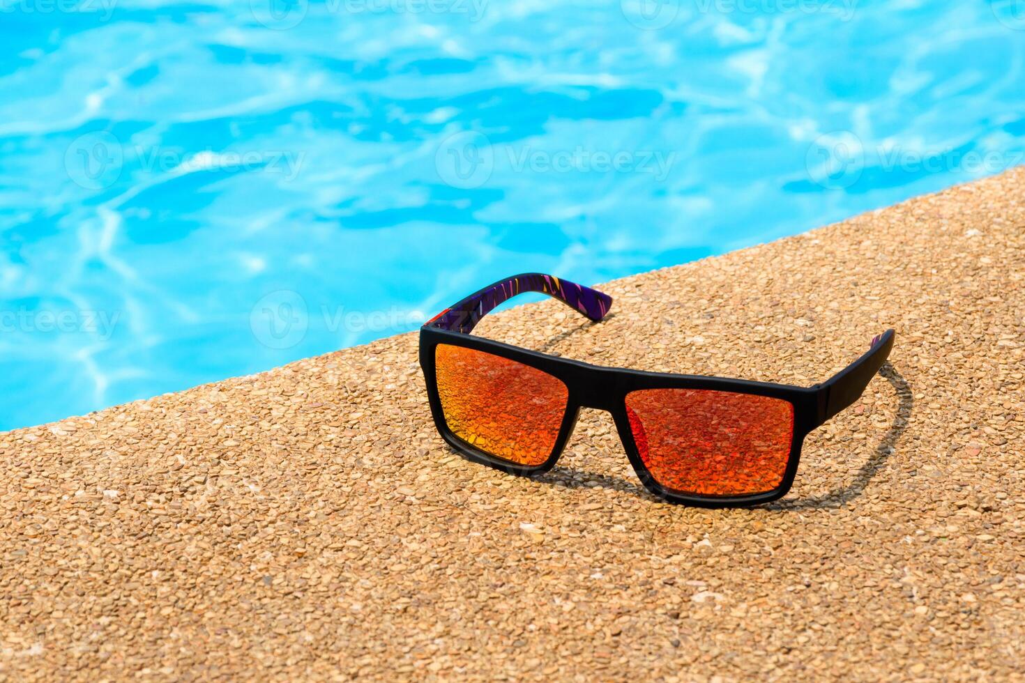 Cool sunglasses on the gravel poolside floor at swimming pool for summer vacation and holiday summer concept, Black sunglasses with orange lenses on tropical season in sunny day. photo