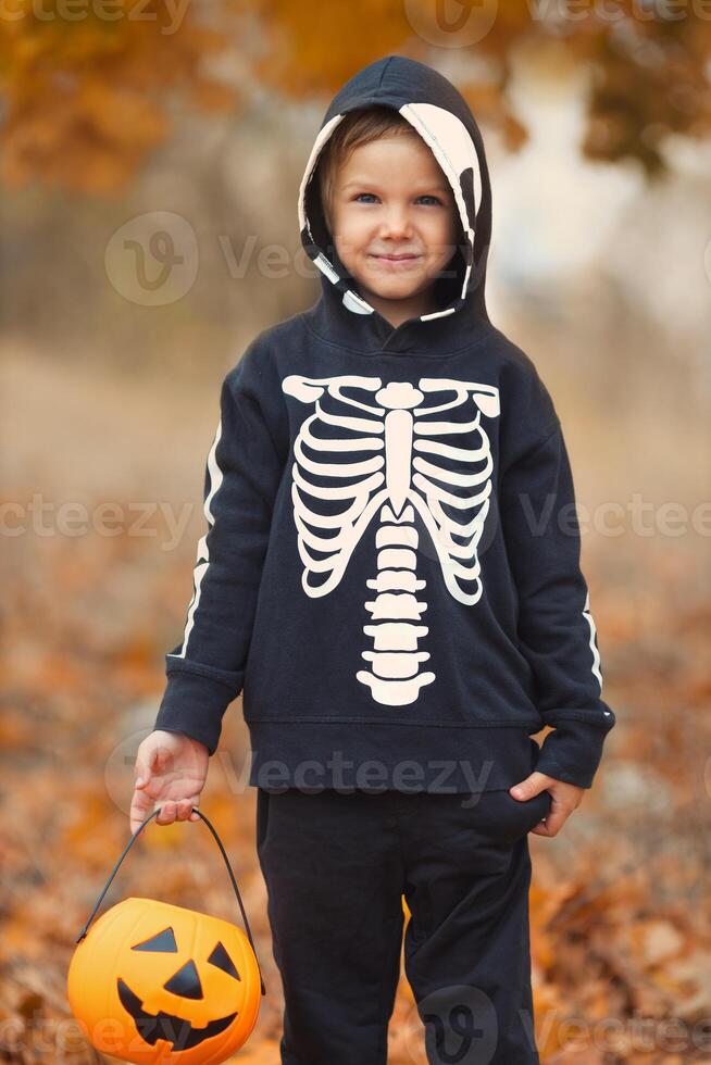 Portrait of funny little boy on spooky skeleton costume for halloween walking in fall park with many orange leaves. Beautiful kid celebrate all saint day with pumpkin basket for trick or treat game. photo