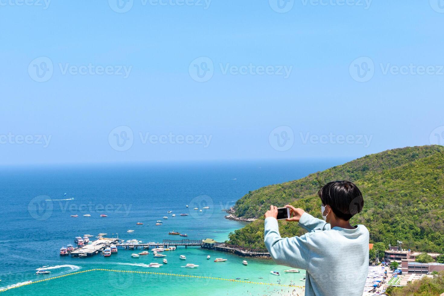 Man holding a smartphone is taking a photo of the view of a tropical beach and an island in the sea on a mountaintop. White sandy beach view Turquoise water at Koh Larn Islands, Thailand.
