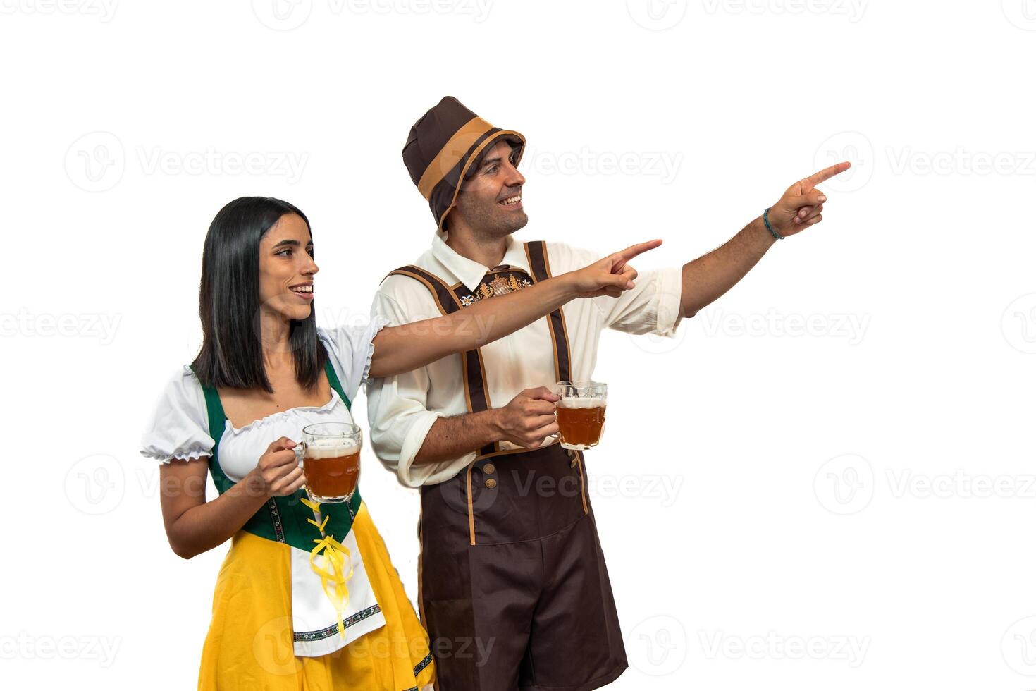 Smiling young Bavarian couple pointing to the right while holding beer glasses at Oktoberfest photo
