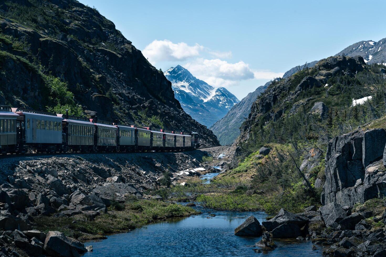 Train cars traveling through an Alaskan mountain pass along a stream of water with snowy peaks in the background and a blue sky with white clouds in the distance. photo