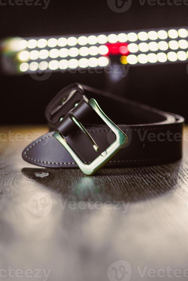 This is a stylish black leather belt featuring a shiny buckle, elegantly displayed on a wooden surface under gentle, soft lighting, showcasing its aesthetic appeal and refined design photo