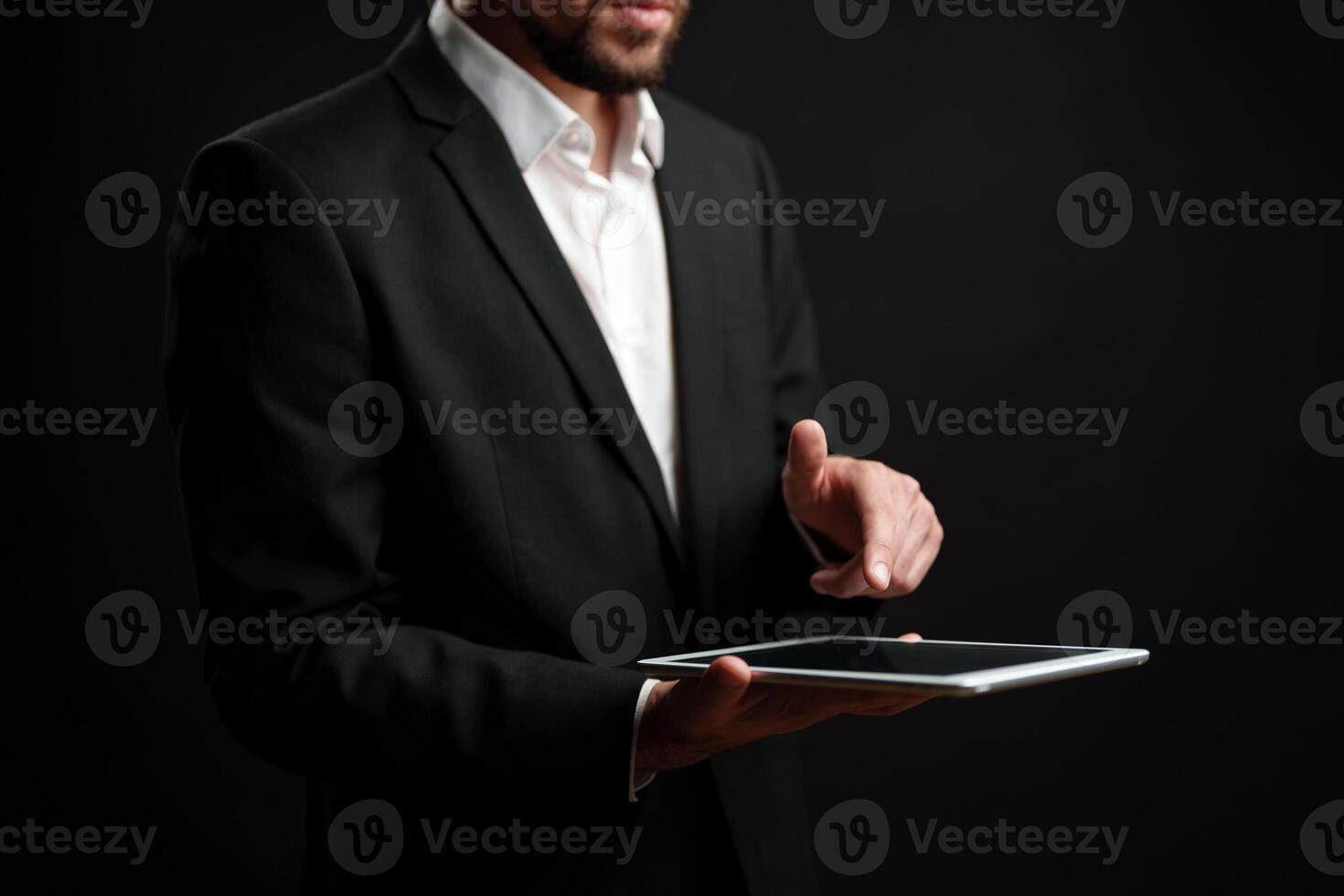 Professional man in suit using tablet while standing against dark background indoors photo